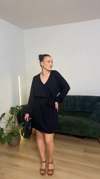 Fekete, hosszú ujjú ruha V-nyakkivágással és megkötős derékkal. Barna platform szandállal viselve. A háttérben zöld kanapé és növény látható.