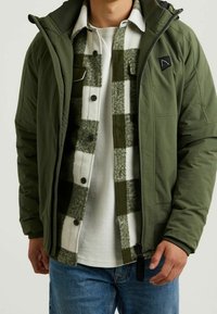 Zelená izolační bunda přes bílou košili a kostkovanou flanelovou košili. Obsahuje zip, boční kapsy a nášivku s logem na hrudi.