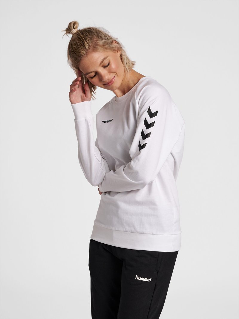 Vrouw met blond haar in een knot, draagt een witte lange mouwen top met zwarte chevrons op de mouwen en zwarte broek, staand tegen een witte achtergrond.