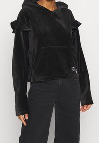 Svart sammetscroppad hoodie med volangdetaljer på ärmarna, framficka och broderad hjärtdesign. Mjuk textur och avslappnad passform.