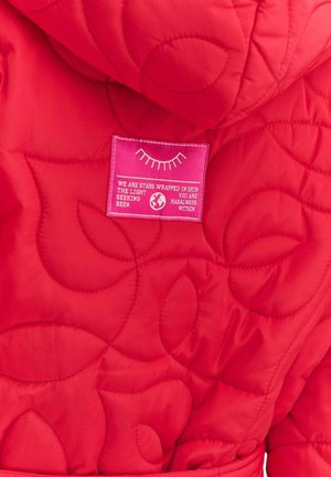 Červená prešívaná bunda s kapucňou a výraznou ružovou štítkom s textom. Tkanina má hladkú, jemnú štruktúru s jemným vzorom.