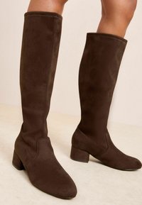 Botas marrones hasta la rodilla hechas de suave ante, con un tacón cuadrado y un diseño liso y sin costuras, sin sujetadores visibles.