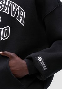 Černá mikina s bílým textem "HIGHVR" a "OCT10." Má ribbované manžety a tkanou nášivku na rukávu. Měkká textura, pohodlný design.