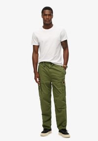 Olijfgroene cargobroek met zijzakken, een verstelbare tailleband en een witte t-shirt met korte mouwen. Zwarte schoenen maken de look compleet.