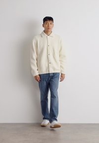 Cremefarvet fleecejakke med krave, knaplukning og sidelommer, parret med lyseblå jeans og hvide sneakers.