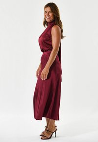 Burgundi satino suknelė su aukštu kaklu ir laisvai krentančia sijonu, su šoniniu įpjovimu ir akcentais, derinama su juostelėmis aukštakulniais.