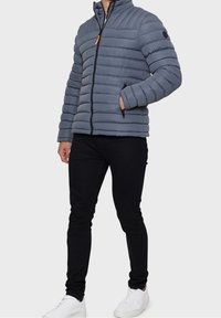 Giacca a vento grigia con design a zigzag orizzontale, chiusura a zip, colletto rialzato e tasche laterali. Indossata con jeans neri e sneakers bianche.