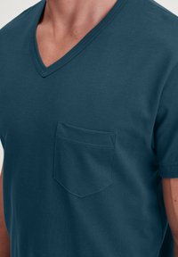 T-shirt pour homme à manches courtes en V de couleur vert foncé, en coton doux. Comprend une poche poitrine avec un ourlet plat et des détails de couture subtils.