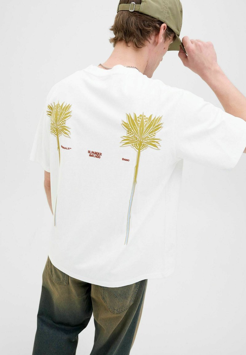 Hvid t-shirt med gule botaniske print på ryggen, med teksten "EL PARQUE del TAMA," iført grønne, falmede jeans.