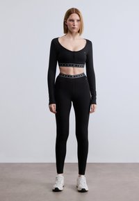Μαύρο crop top με μακριά μανίκια, στρογγυλή λαιμόκοψη και λεπτομέρεια με δύο κουμπιά, σε συνδυασμό με μαύρο κολάν ψηλόμεσο με λογότυπο στη ζώνη.