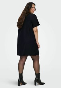 Melnā T-krekla kleita ar īsām piedurknēm, kurai ir teksturētas akcenti. Modelis valkā melnus potīšu zābakus un caurspīdīgas zeķes, nedaudz pagriežoties atpakaļ.