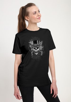 Kvinde med hestehale bærer en sort Guns N' Roses t-shirt med en kranie med høj hat og krydsede pistoler, kigger let opad og smiler.