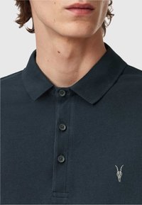 Dunkelblaues Polohemd aus strukturiertem Stoff, mit drei schwarzen Knöpfen und einem kleinen Ziegenschädel-Logo, das auf der linken Brust gestickt ist.