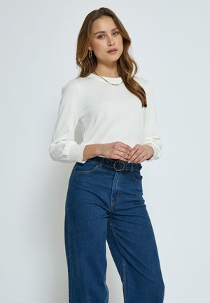 Haut à manches longues blanc avec des poignets côtelés, associé à un jean en denim bleu taille haute avec une boucle noire ronde. Design simple et classique.