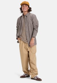 Grijze flanellen shirt over een gestreept t-shirt, beige baggy broek en bruine sneakers met witte strepen. Accessoires zijn een tan pet.