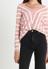 Roze en witte gestreepte trui met een V-hals en brede mouwen, gemaakt van een zacht breigoed, gecombineerd met hoge zwarte jeans.