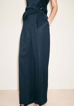 Osoba nosząca czarne, szerokie spodnie z wysokim stanem, ręce w kieszeniach na jednolitym jasnym tle.