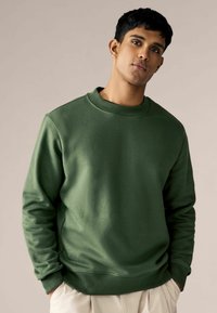 Groen sweatshirt met een geribbelde ronde hals, lange mouwen en een gladde textuur. Heeft een ontspannen pasvorm en is gecombineerd met lichte broeken.