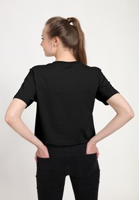Černé tričko s krátkým rukávem a kulatým výstřihem, vyrobené z měkké látky. Model ho nosí s tmavými džíny a má ruce v zadních kapsách.