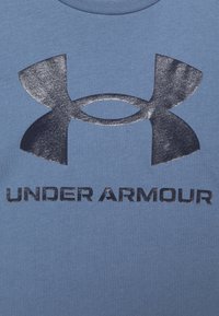 Modrá bavlněná tričko s metalickou tmavě modrou logem Under Armour a textem v tučném, moderním písmu. Krátké rukávy a kulatý výstřih.
