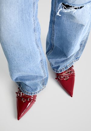 Lysblå ripped jeans båret med spidse, glinsende røde sko med flere spænder på en hvid baggrund.