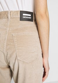 Beiga kordfläckiga byxor med en texturerad yta, med bakfickor och en svart märkesetikett med vit logotyp på midjebandet.