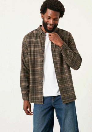 Braun karierte Hemdjacke aus strukturiertem Stoff, über einem weißen T-Shirt getragen. Lockerer Schnitt mit langen Ärmeln, kombiniert mit blauen Denim-Hosen.