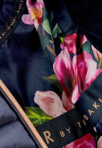 Kwiatowy materiał z różowymi i białymi kwiatami wewnątrz ciemnej kurtki z złotym zamkiem błyskawicznym oraz napisem "TED BAKER" na czarnej wstążce.
