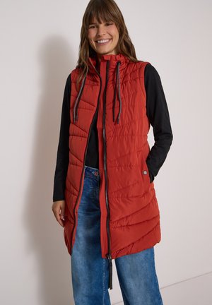 Femme souriante portant une veste matelassée rouge sans manches avec des manches longues noires et un jean bleu sur un fond clair uni.