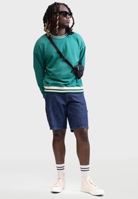 Groene sweatshirt met geribde kraag en zoom, denim shorts, zwarte schoudertas, witte sokken met zwarte strepen, beige hoge sneakers.