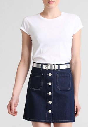 Femme portant un t-shirt blanc aux manches retroussées, rentré dans une jupe en denim bleu foncé à boutons sur le devant, avec des poches plaquées et une ceinture argentée.