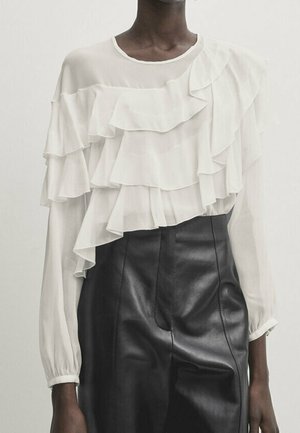 Personne portant un chemisier blanc à volants, manches longues, rentré dans un pantalon en cuir noir taille haute, sur un fond uni.