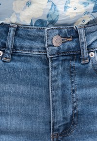 Μέση μπλε τζιν με μεταλλικό κουμπί και θηλιές για ζώνη, σε συνδυασμό με πουκάμισο ανοιχτού μπλε χρώματος με λουλουδάτο μοτίβο, που είναι μπλουκωμένο μέσα.