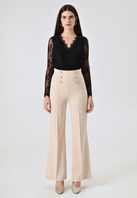 Zwarte kanten top met lange mouwen en een V-hals, gecombineerd met beige hoge taille flare broeken met gouden knoopaccenten.