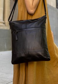 Black leather shoulder bag with a smooth texture, featuring a front pocket and adjustable strap, worn against a tan dress.