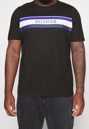 Mann trägt ein schwarzes T-Shirt mit horizontalen weißen, blauen und schwarzen Streifen und dem Schriftzug „HILFIGER“ auf der Brust, kombiniert mit dunklen Jeans.