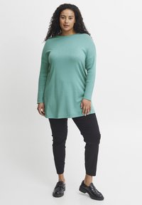Tunica turchese a maniche lunghe con vestibilità rilassata, texture a costine e spacchi laterali, abbinata a pantaloni neri slim-fit e mocassini neri.