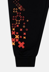 Černý látkový rukáv s červenými, oranžovými a žlutými vzory ve tvaru X a kruhů shlukujícími se do diagonálního designu blízko manžety.