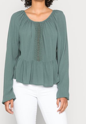 Vrouw draagt een groene blouse met lange mouwen en kantdetail aan de voorkant en een witte broek, staand tegen een effen achtergrond.
