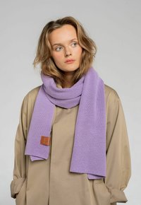 Fioletowy dziergany szalik wykonany z miękkiego materiału, z teksturowanym splotem i brązową skórzaną metką. Ułożony i stylizowany z beżowym płaszczem.