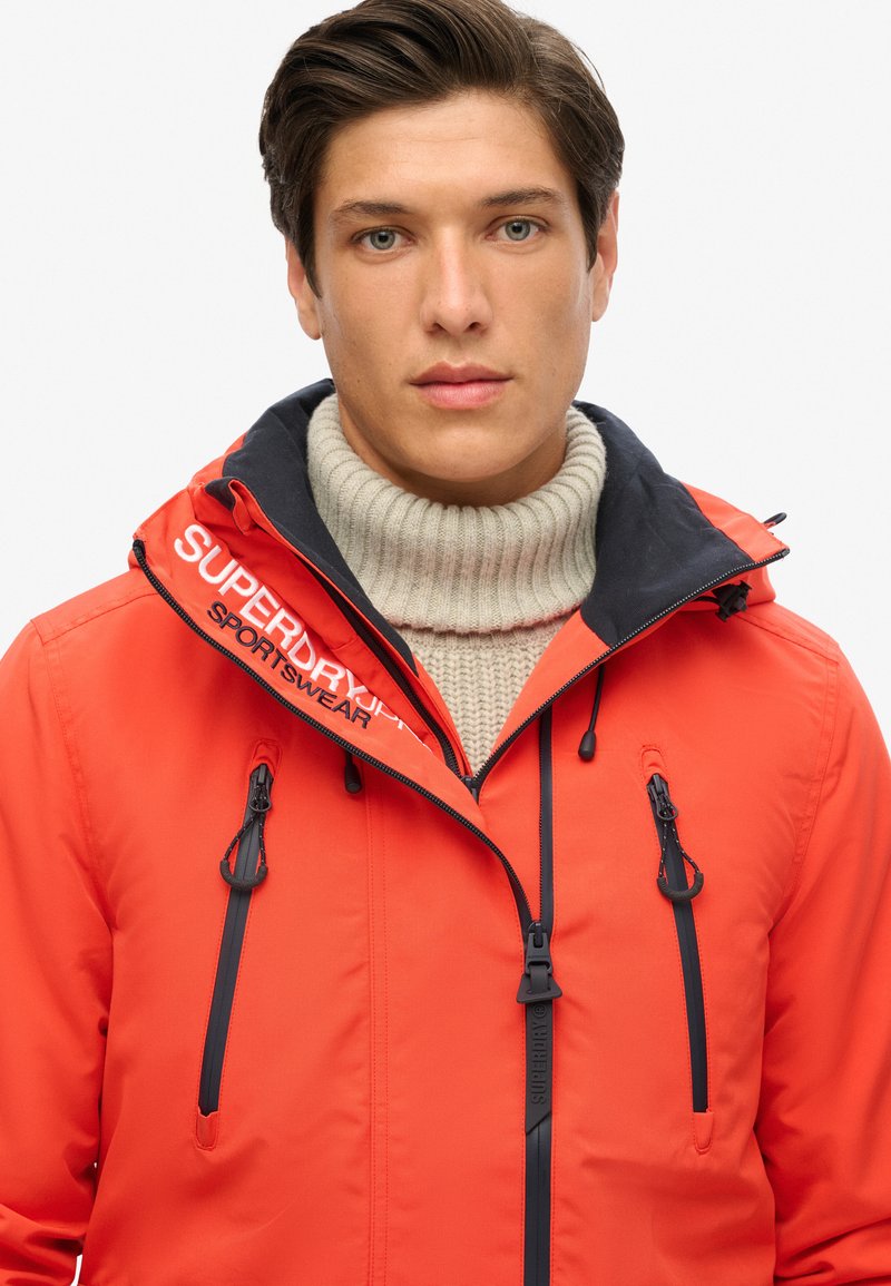 Chaqueta impermeable roja con un collar negro, que cuenta con un cierre de cremallera y cordones ajustables. Logotipo en el hombro izquierdo; dos bolsillos frontales.