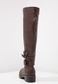 Bota de cuero marrón hasta la rodilla con dos correas ajustables con hebilla y una superficie texturizada; tacón bloque robusto para mayor estabilidad.
