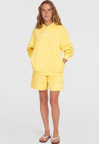 Felpa gialla con tasche a marsupio e testo bianco, abbinata a pantaloni corti coordinati. Materiale in cotone morbido, vestibilità rilassata e senza motivi visibili.