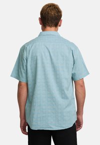 Camisa de manga corta, de color azul claro, con un patrón geométrico blanco, que presenta un cuello, corte recto y un bolsillo de parche en la parte posterior.
