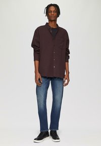 Chemise boutonnée bordeaux foncé avec une poche, superposée sur un haut noir, assortie à un jean bleu et des baskets noires avec des semelles blanches.