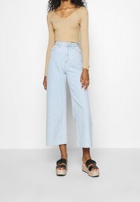 Kobieta ubrana w beżową, prążkowaną bluzkę z długim rękawem, jasnoniebieskie, wysokie dżinsy z szerokimi nogawkami oraz czarne sandały na platformie z klamrami.