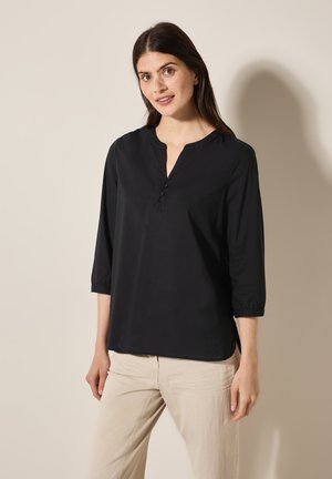 Schwarze Bluse mit Dreiviertelärmeln, Rundhalsausschnitt und Knopfleiste. Weicher Stoff mit lockerer Passform. Kombiniert mit hellbeigen Hosen.
