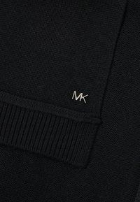 Czarna dzianina o ryflowanej fakturze. Zawiera małe srebrne logo "MK" na krawędzi, podkreślające design.