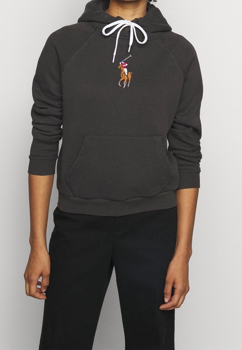 Person trägt einen dunkelgrauen Kapuzenpullover mit weißen Kordeln und einem Polo-Spieler-Logo auf der Brust, kombiniert mit schwarzen Hosen.