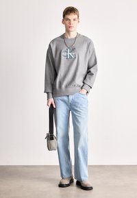 Mladý muž stojí v šedé mikině Calvin Klein, světlých modrých džínách, béžových botách, se stříbrným náhrdelníkem a drží malou šedou tašku s černým popruhem.